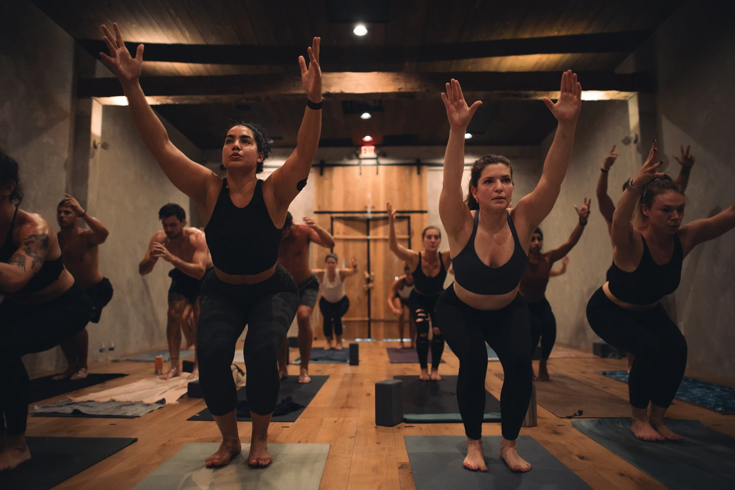 A group reaches upward from bent-knee stances beneath the studio’s reclaimed beams.