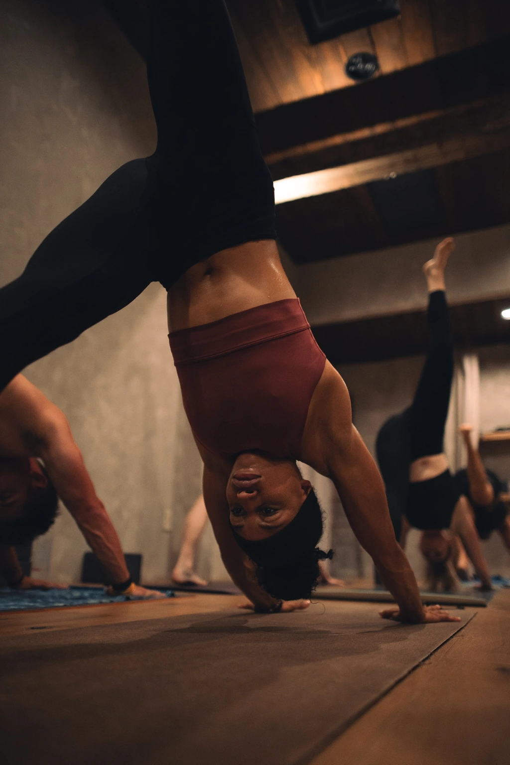 A practitioner balances in an inverted pose, with her hands on the mat and other students practicing beneath the reclaimed studio beams.