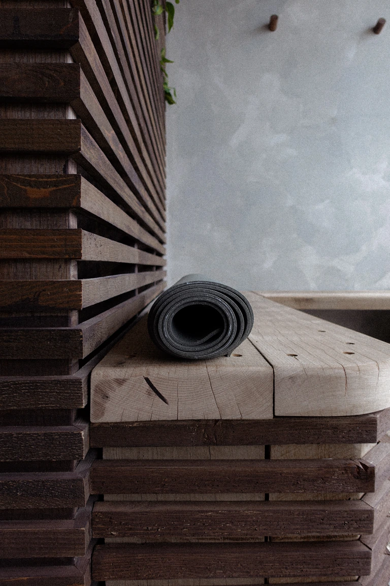 A rolled black yoga mat on a raw timber bench beside dark wooden slats and a limewashed wall.