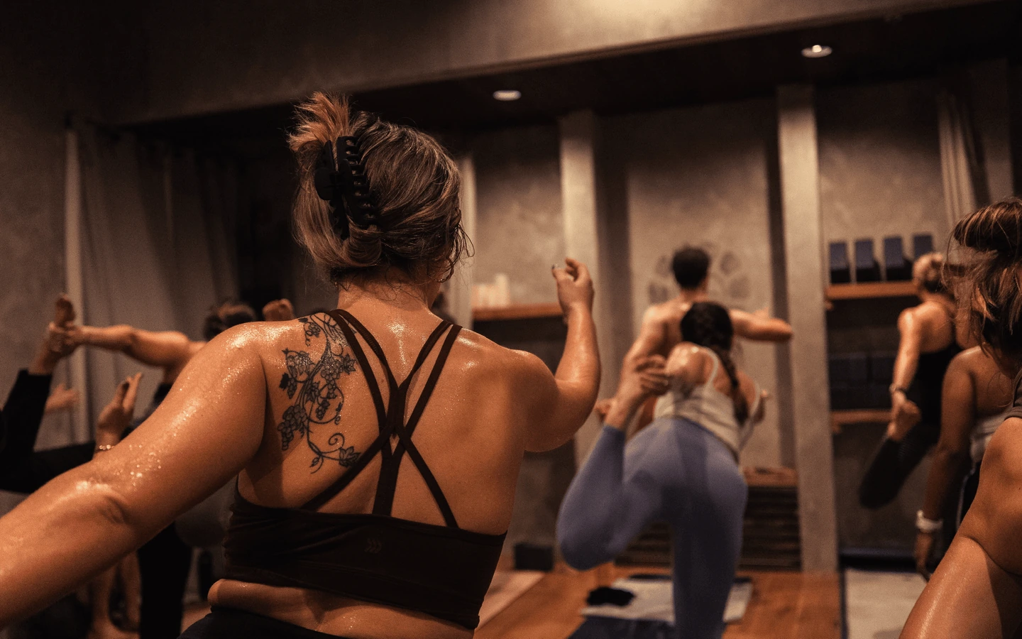 A tattooed practitioner in a black crisscross top balances with the class in the REWILD YOGA studio.