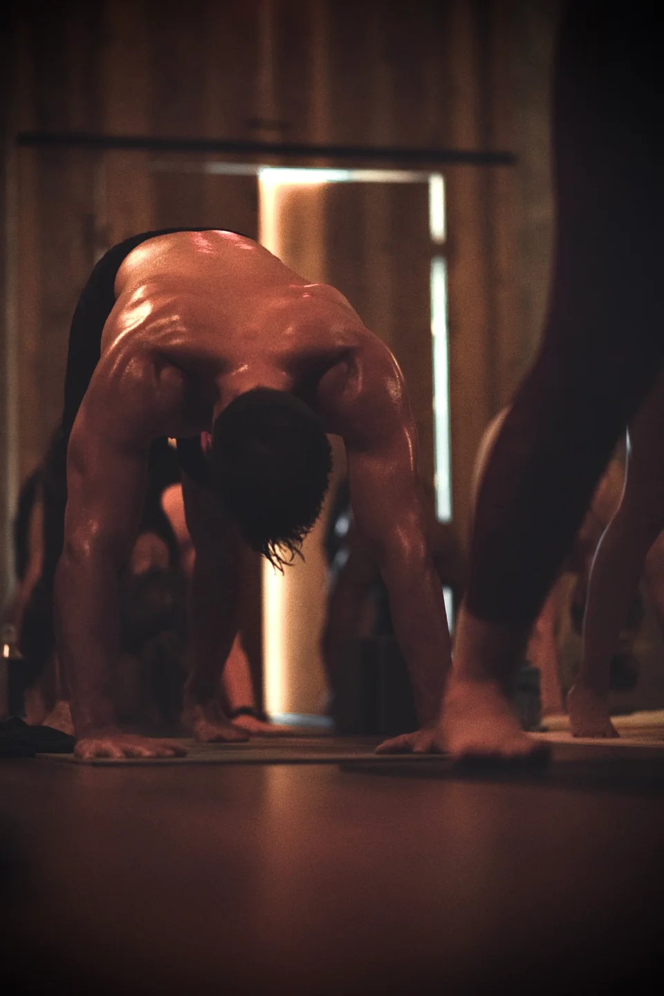 A practitioner in a forward-loaded posture, sweat catching the light across his back and arms.